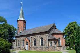 Kirche von rechts, vorn 2 Steige und Laterne links, grüne Eichen beidseitig, im Hintergrund rechts Gutsscheune